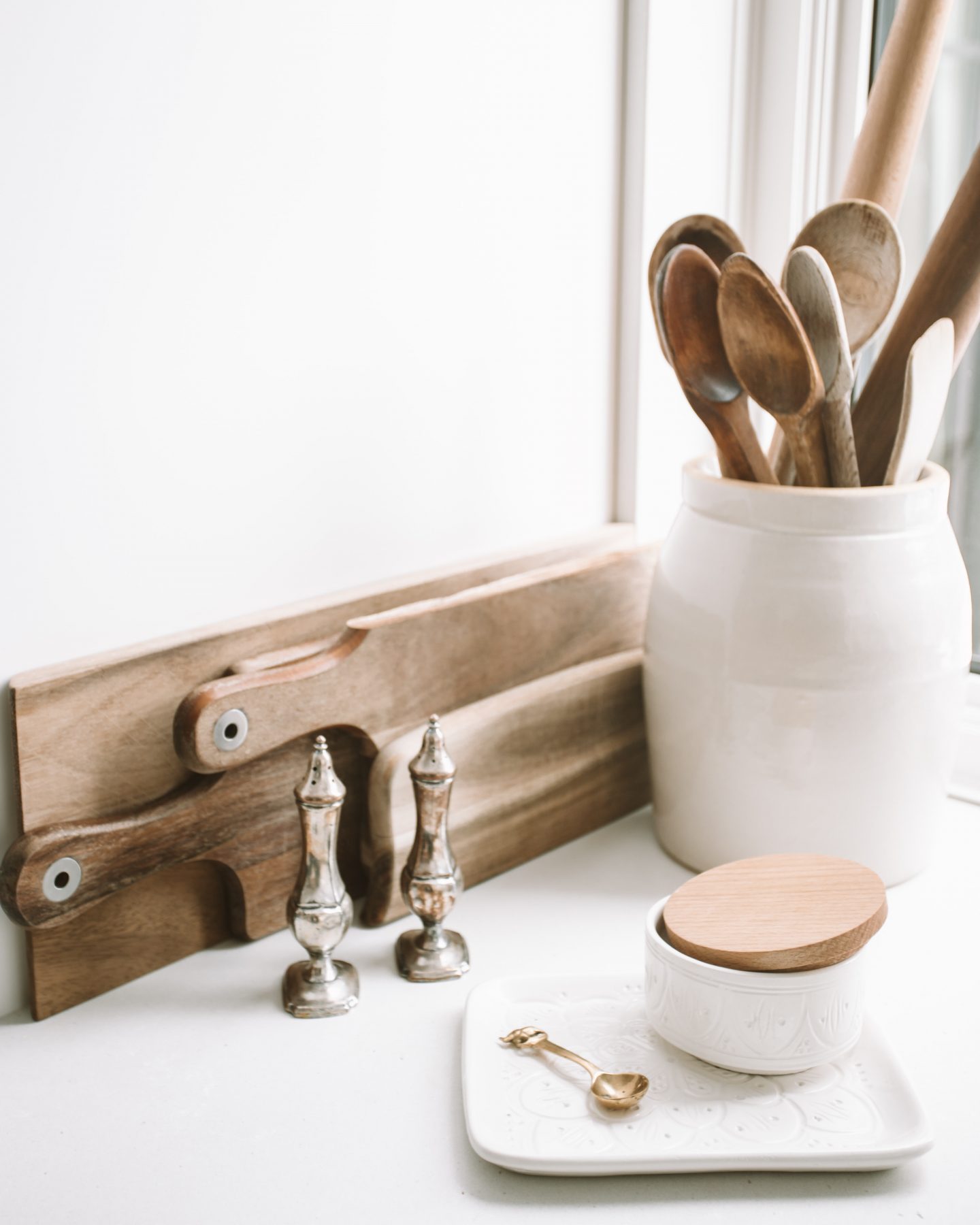 Kitchen counter with crock and wooden spoons. Silver salt and pepper shakers. Cutting boards leaning against the wall.