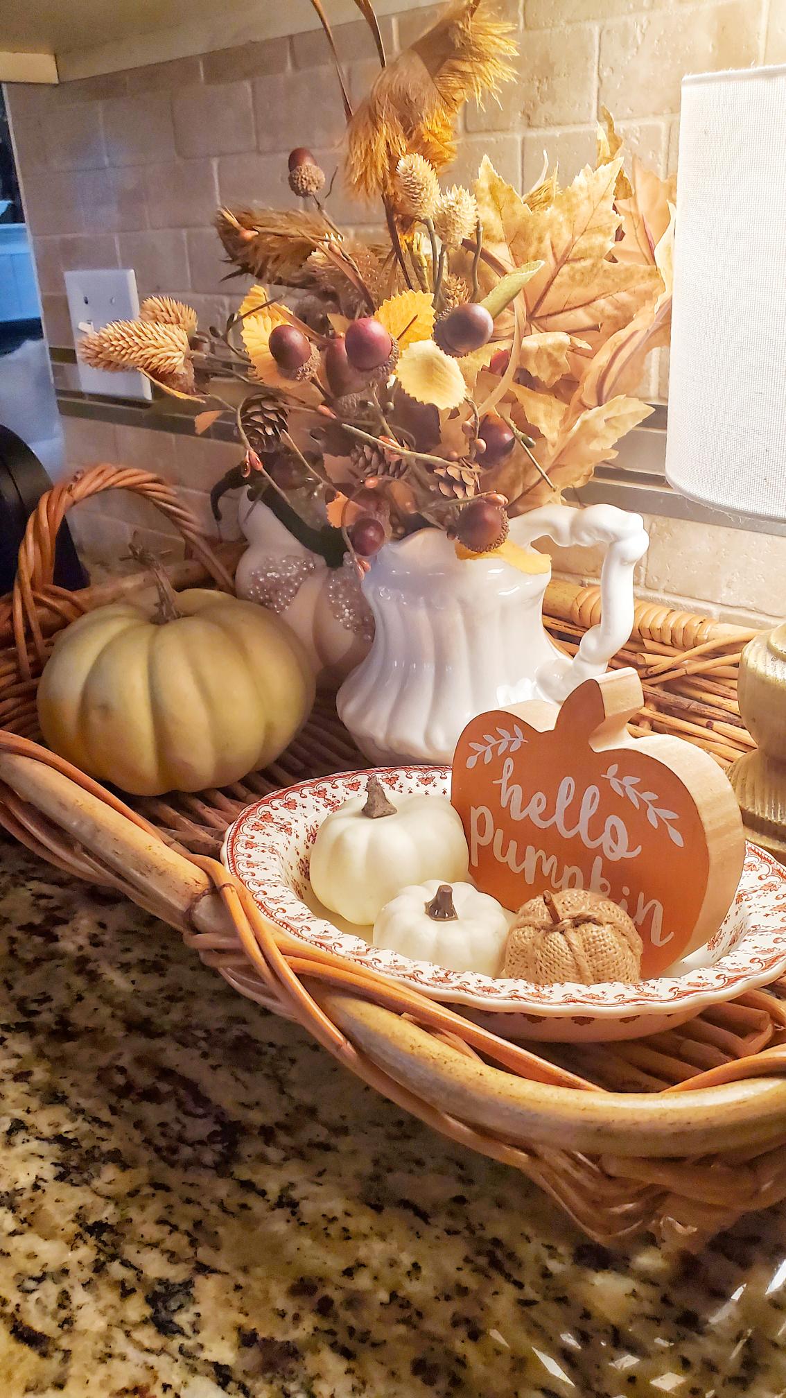 In a basket on top of our kitchen counter is a bouquet of flowers, pumpkins and a "hello fall" sign.
